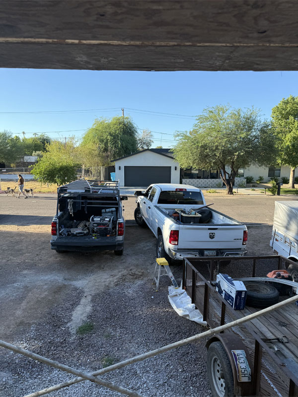 Two work trucks parked in a residential area, one with a ladder rack and tools visible, alongside a trailer, with a house and trees in the background, representing AZ Garage Door Pros LLC's service vehicles.