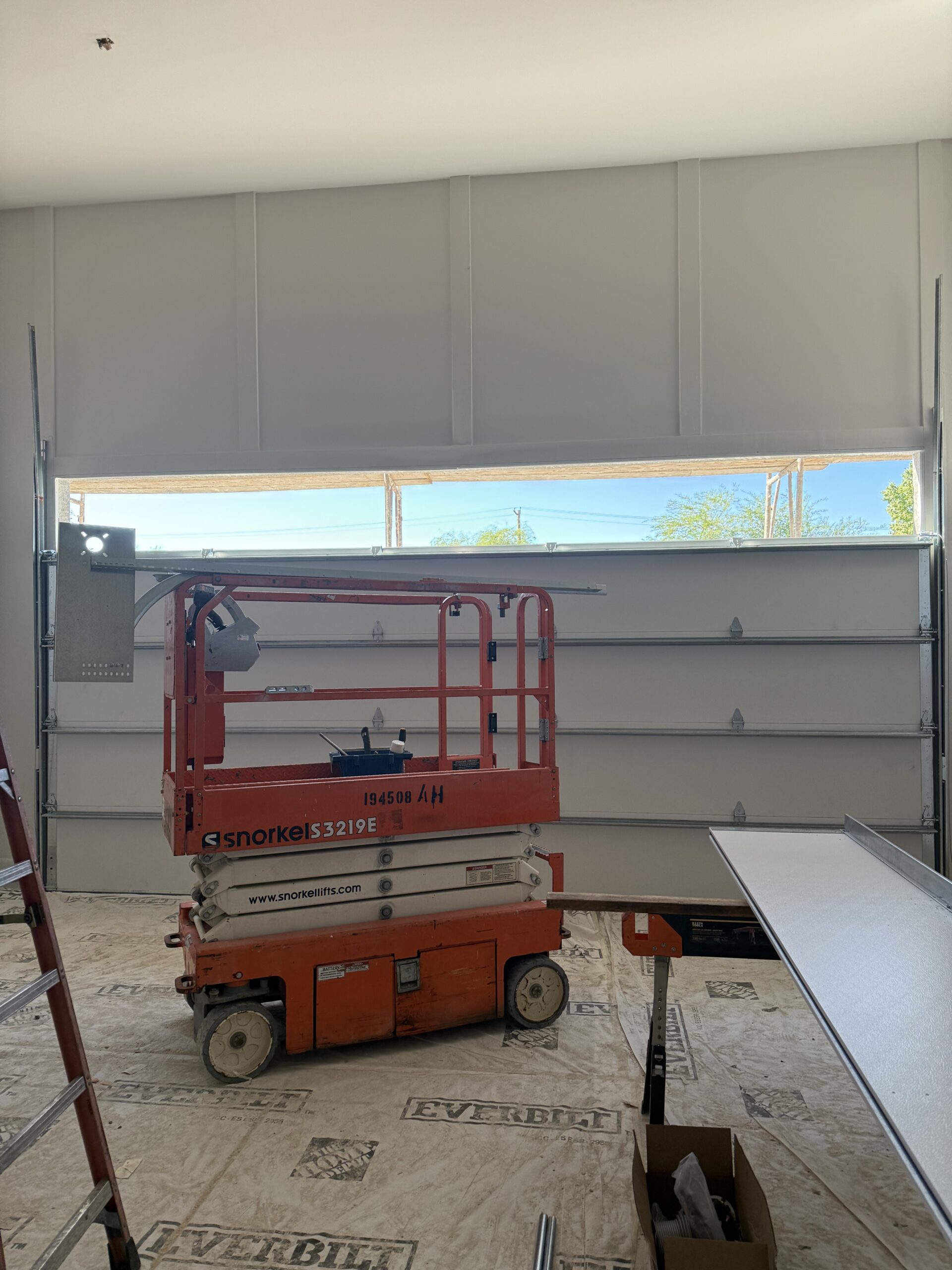 Interior view of garage under construction featuring a Snorkel lift, exposed garage door tracks, and a ladder on the floor, illustrating the installation process for garage door services by AZ Garage Door Pros LLC.