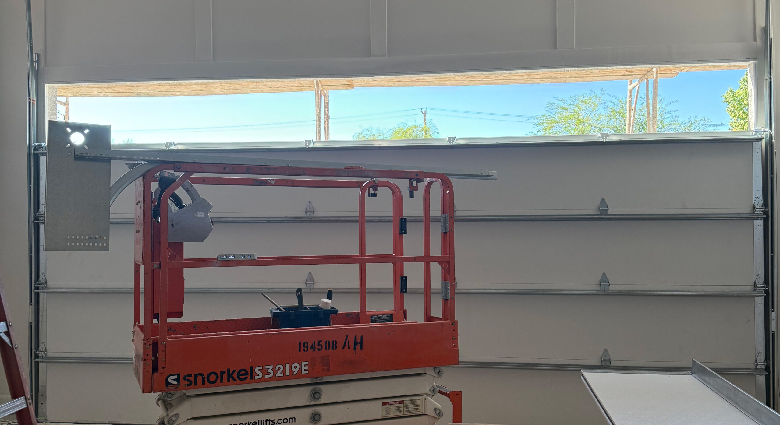 Scissor lift positioned inside a garage, with a view of the open garage door and clear blue sky, illustrating garage door installation or repair services by AZ Garage Door Pros LLC.
