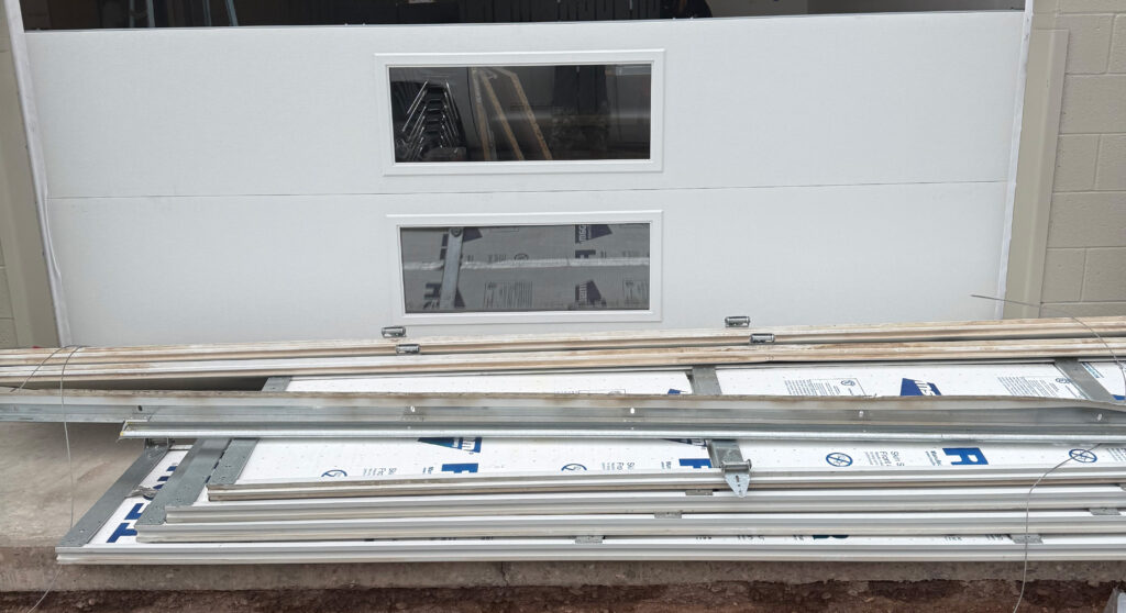 Garage door components and tracks stacked in front of a white garage door, illustrating off-track repair services by AZ Garage Door Pros LLC.