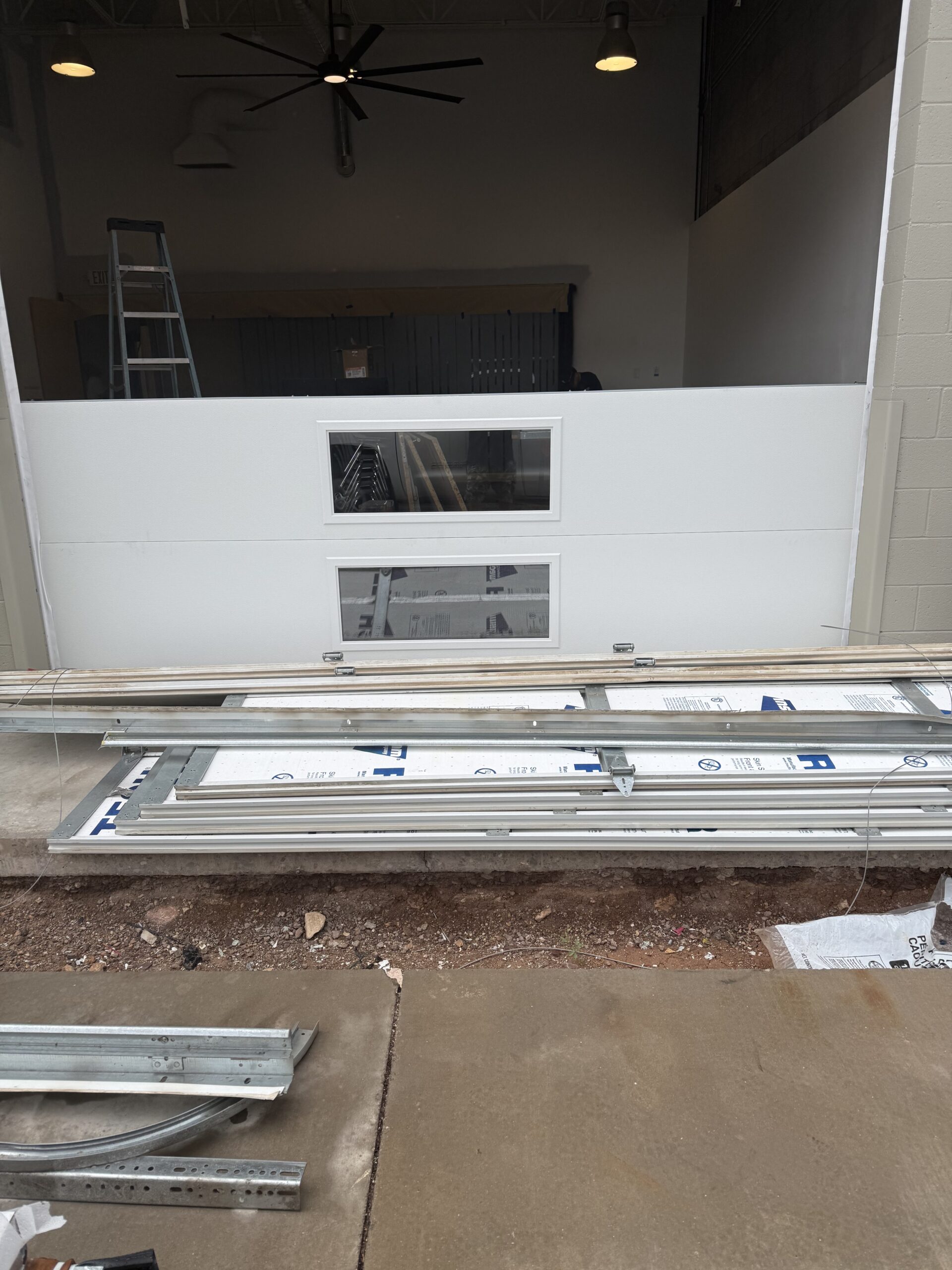 Garage door installation in progress, featuring a partially installed white garage door with windows, a ladder, and various metal components on the ground, showcasing the reliable service of AZ Garage Door Pros LLC.