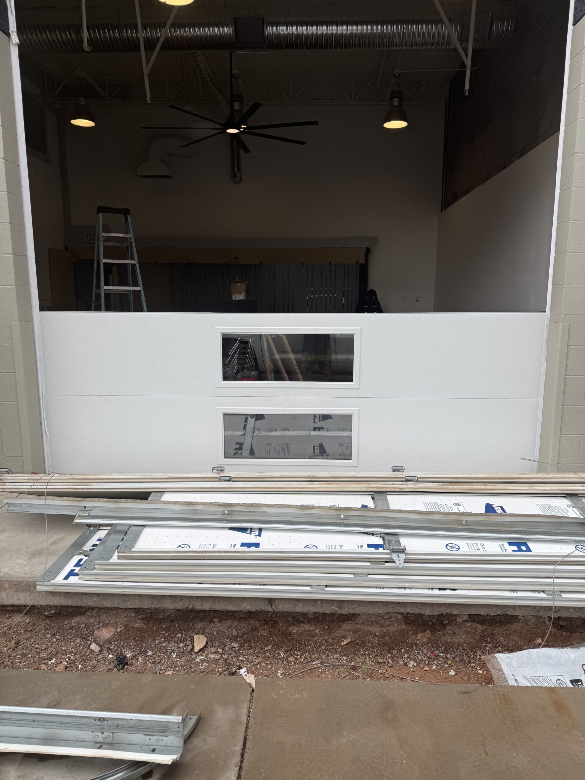 Garage door installation in progress, featuring a white garage door with a window, tools and materials on the ground, and a ladder in a well-lit workspace.
