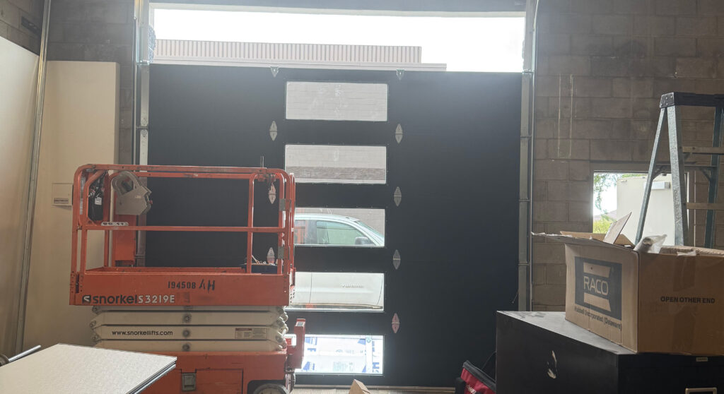 Garage door repair scene featuring a partially opened black garage door with windows, a Snorkel lift positioned in front, and a cluttered workspace in a commercial setting.