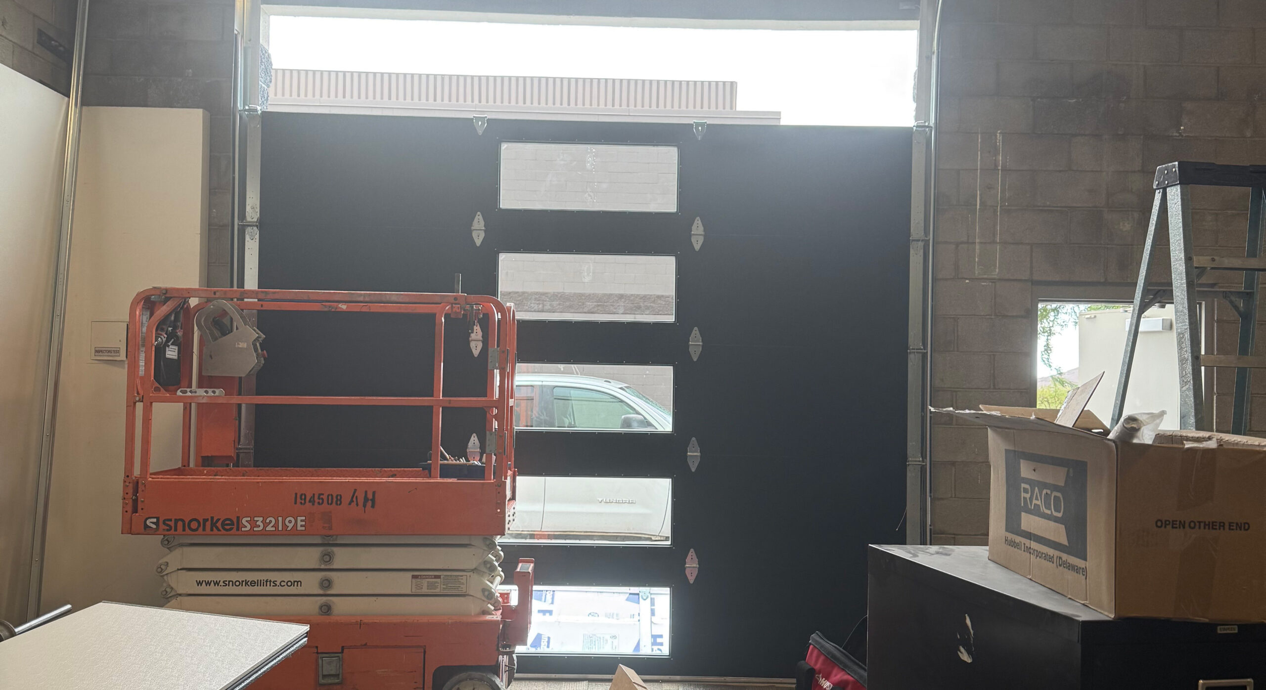 Garage door installation in progress with a black garage door featuring windows, orange lift equipment, and workspace clutter.