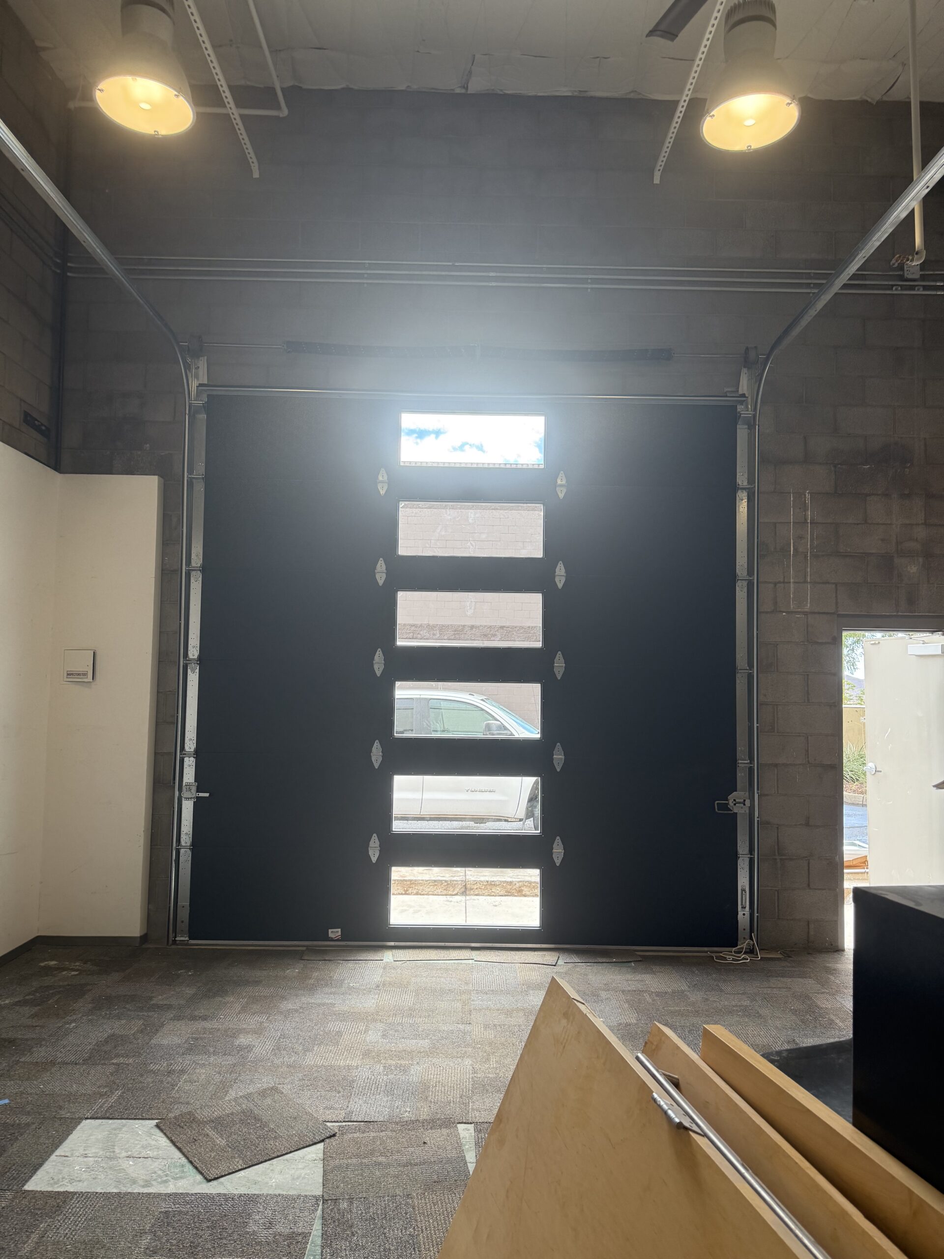 Garage door installation in a commercial space, featuring a modern black garage door with windows, illuminated interior, and unfinished flooring.